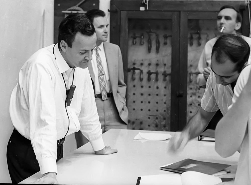 Richard Feynman leaning over a table while wearing the lecture microphone, with several colleagues around him.